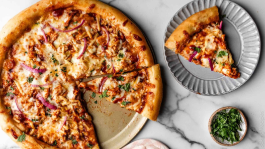 Close-up of a homemade BBQ chicken pizza topped with shredded chicken in tangy sauce, smoked gouda, mozzarella, red onion, and fresh cilantro.