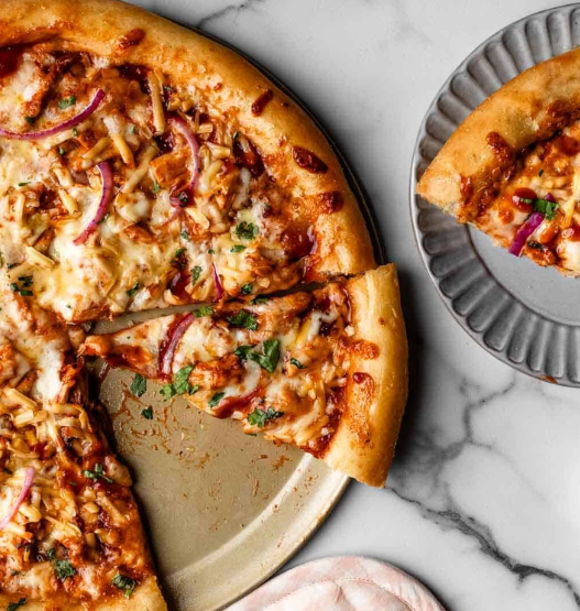 Close-up of a homemade BBQ chicken pizza topped with shredded chicken in tangy sauce, smoked gouda, mozzarella, red onion, and fresh cilantro.