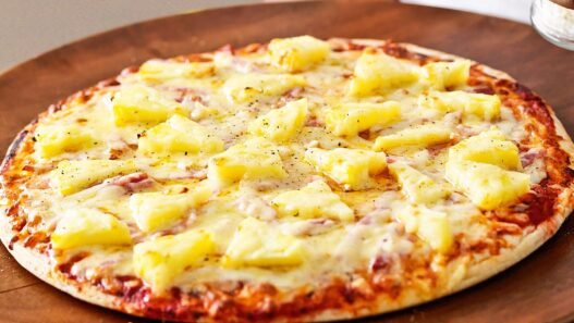 Chef at Lupa Pizza in Norwich holding a Hawaiian pizza priced at £100, created as a satirical response to the pineapple-on-pizza debate.