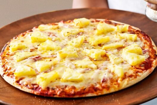 Chef at Lupa Pizza in Norwich holding a Hawaiian pizza priced at £100, created as a satirical response to the pineapple-on-pizza debate.